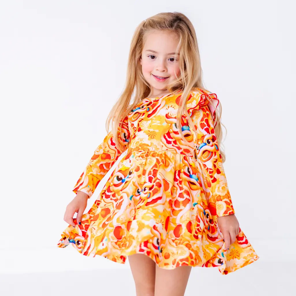 A child twirling with a soft smile and the skirt held out, in a long sleeve twirl dress with flutter ruffles at the shoulders in a golden yellow print of lion dance heads and red lanterns.