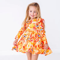 A child twirling with a soft smile and the skirt held out, in a long sleeve twirl dress with flutter ruffles at the shoulders in a golden yellow print of lion dance heads and red lanterns.