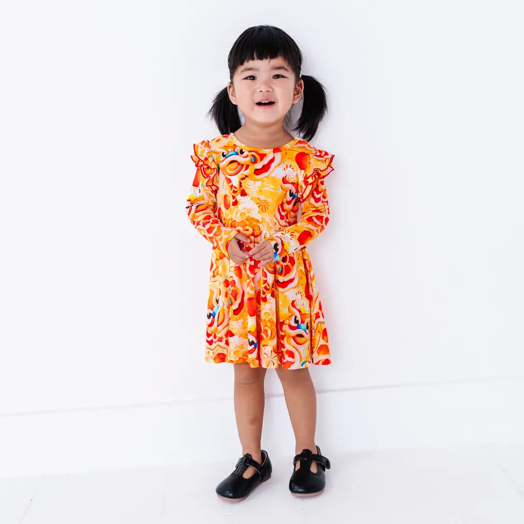 A toddler with pigtails smiling widely with hands clasped, in a long sleeve twirl dress with flutter ruffles at the shoulders in a golden yellow print of lion dance heads and red lanterns.