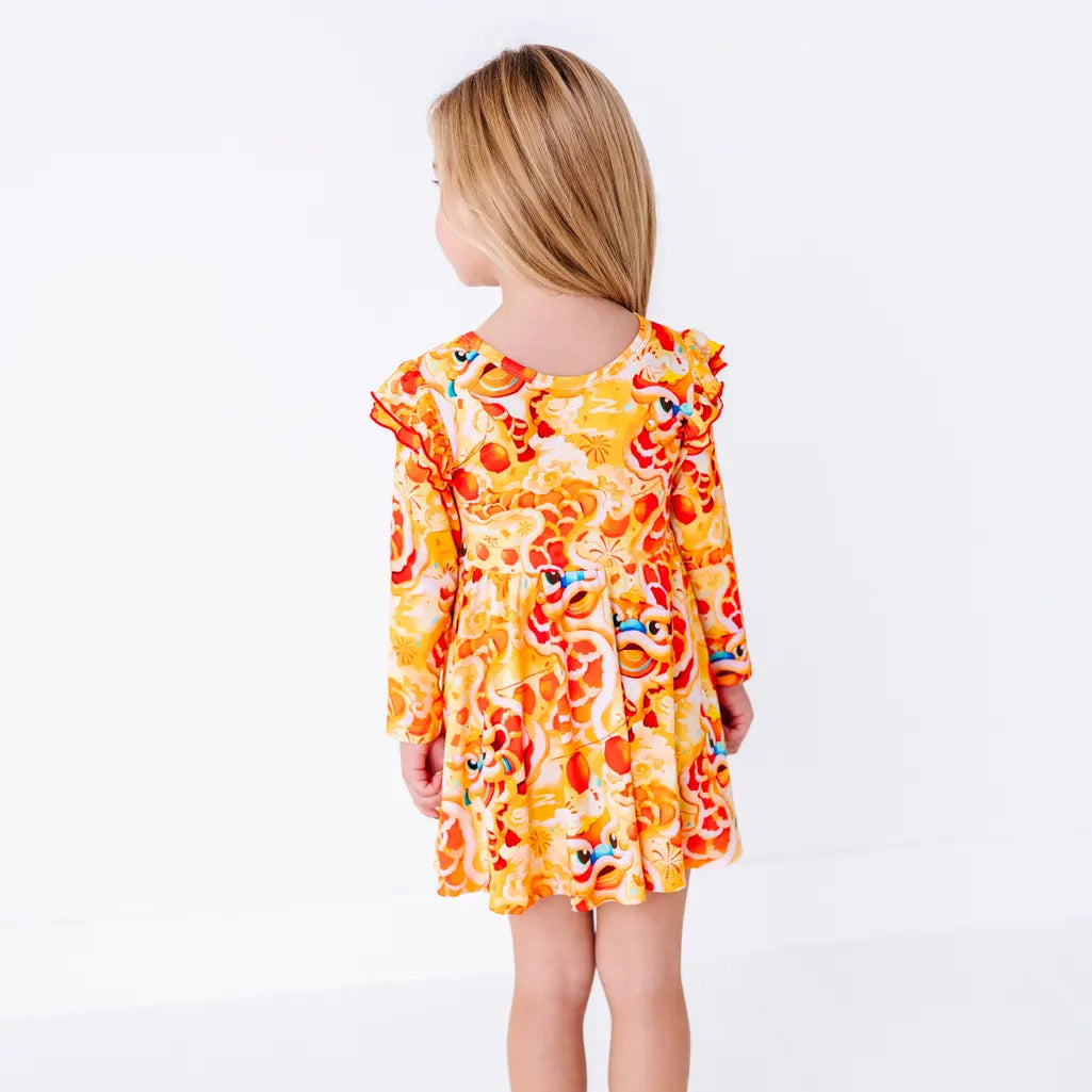 Back view of a child with straight hair and the head turned, in a long sleeve twirl dress with flutter ruffles at the shoulders in a golden yellow print of lion dance heads and red lanterns.