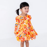 Side view of a toddler with pigtails smiling and looking aside, in a long sleeve twirl dress with flutter ruffles at the shoulders in a golden yellow print of lion dance heads and red lanterns.