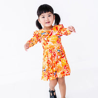 A smiling toddler with pigtails walking with arms out, in a long sleeve twirl dress with flutter ruffles at the shoulders in a golden yellow print of lion dance heads and red lanterns.