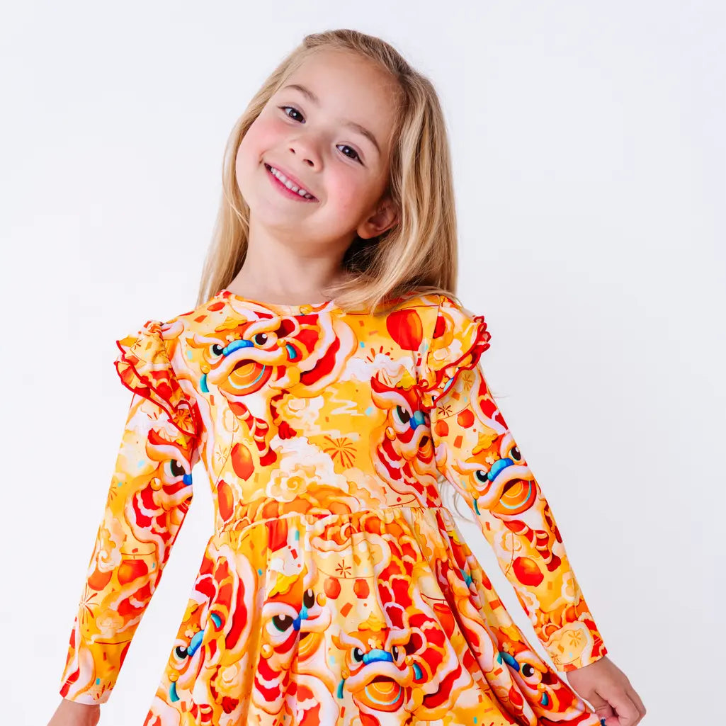 Close up portrait of a child with a big smile and the head tilted, in a long sleeve twirl dress with flutter ruffles at the shoulders in a golden yellow print of lion dance heads and red lanterns.