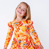 Close up portrait of a child with a big smile and the head tilted, in a long sleeve twirl dress with flutter ruffles at the shoulders in a golden yellow print of lion dance heads and red lanterns.