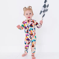 A baby standing in a cream zip-up pajama with a colorful monster truck print, holding a checkered racing flag.