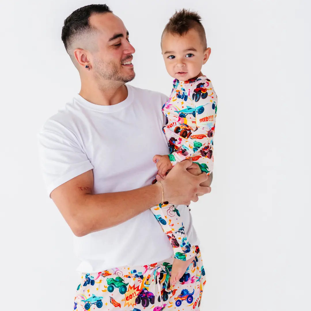 A father in a white tee holding a baby dressed in a cream pajama with a colorful monster truck print.