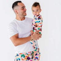 A father in a white tee holding a baby dressed in a cream pajama with a colorful monster truck print.