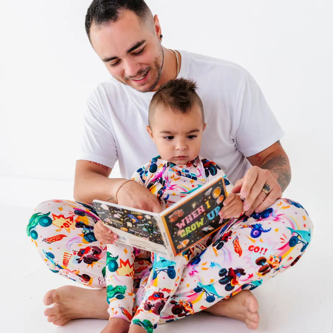 A father in monster truck print pajama pants reading the storybook When I Grow Up with a baby in matching pajamas.