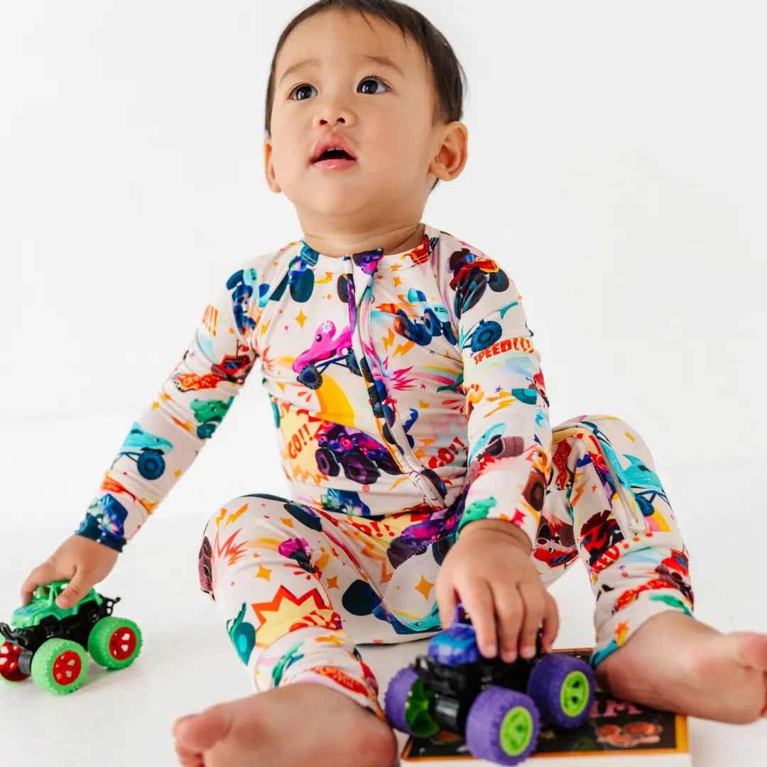 A baby sitting with toy monster trucks while wearing a cream zip-up pajama with a colorful monster truck print.