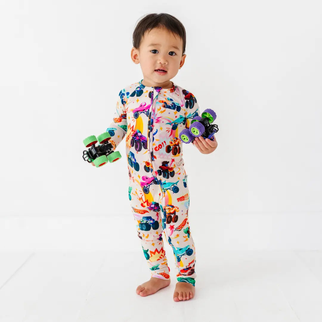 A baby standing and holding two toy monster trucks, wearing a cream zip-up pajama with a colorful monster truck print.