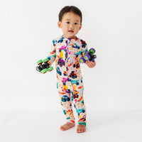 A baby standing and holding two toy monster trucks, wearing a cream zip-up pajama with a colorful monster truck print.