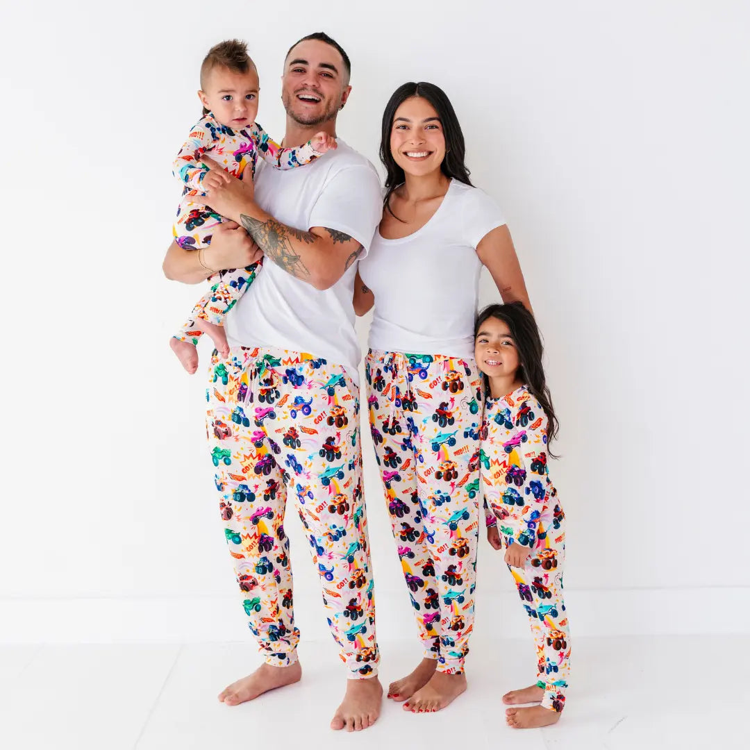 A family in matching monster truck print pajama bottoms and white tops, with a baby and a child, smiling together.