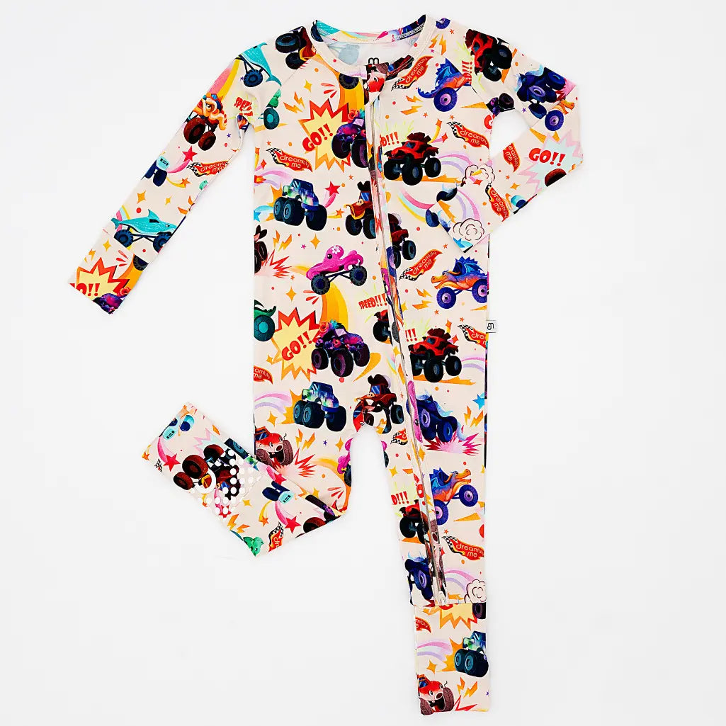 Flat lay of a cream zip-up convertible baby pajama with a colorful monster truck print and comic-style bursts on a white background.