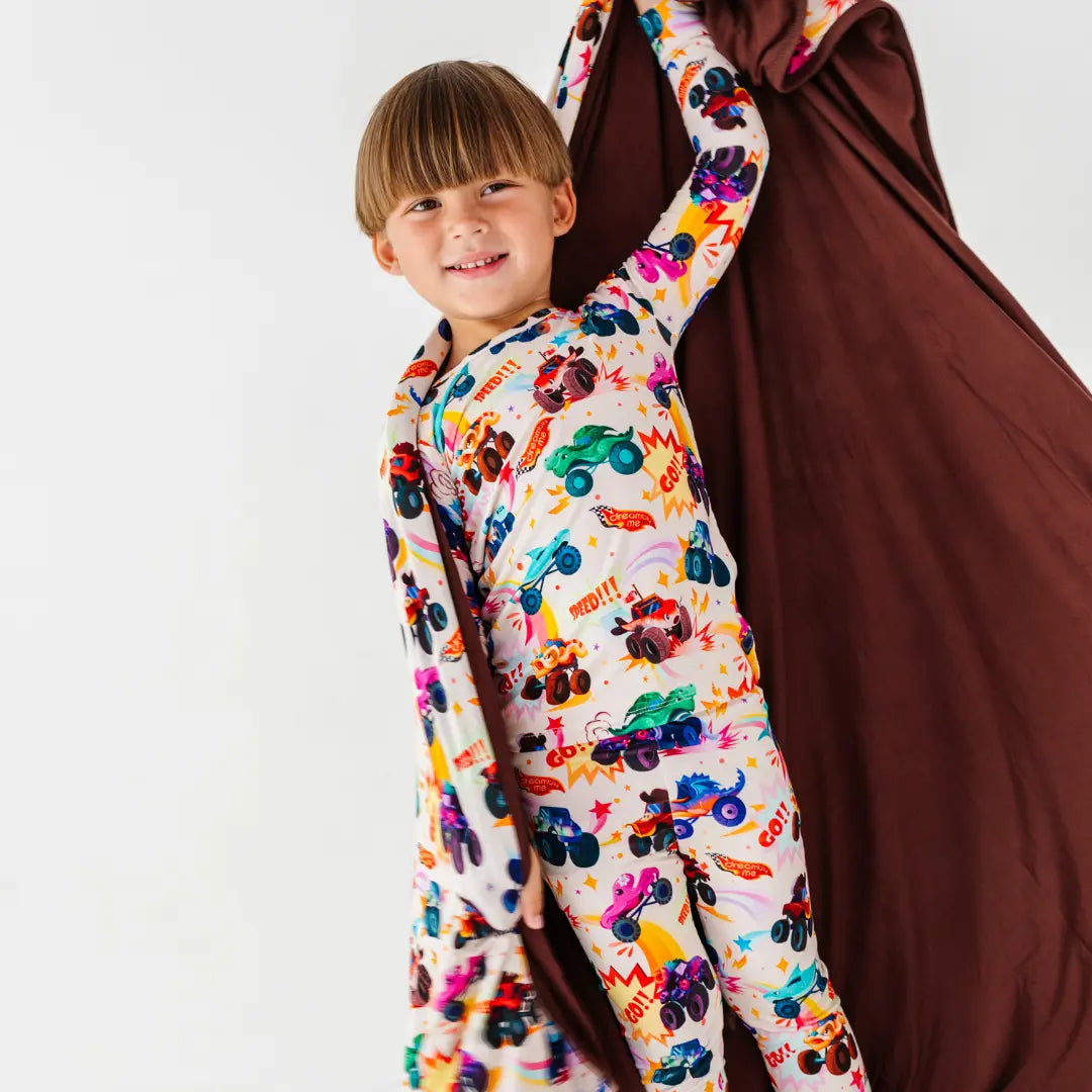 A child in monster truck print pajamas holding up a blanket showing its solid brown reverse side.