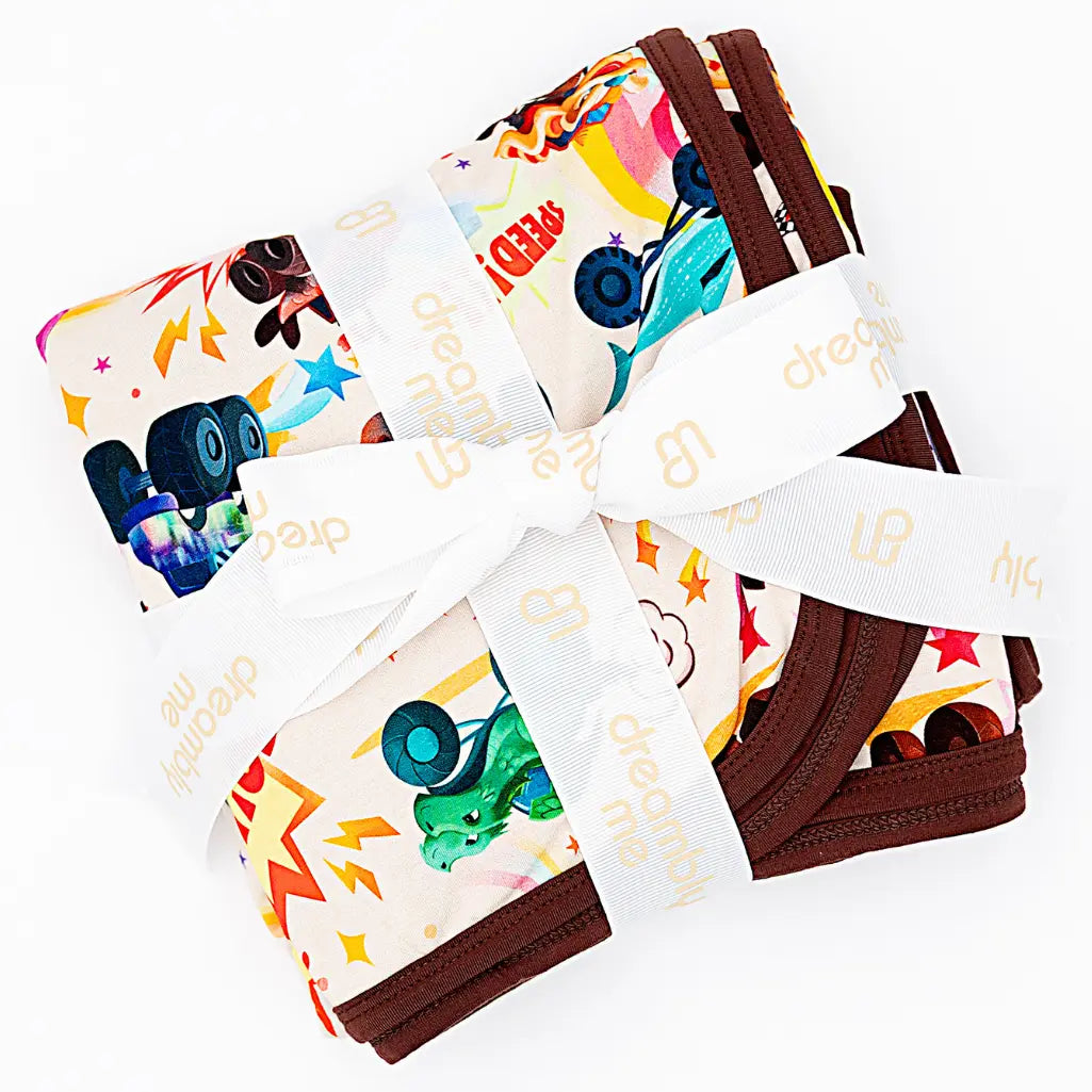 Flat lay of a folded blanket with a colorful monster truck print and brown trim, tied with a white ribbon, on a white background.