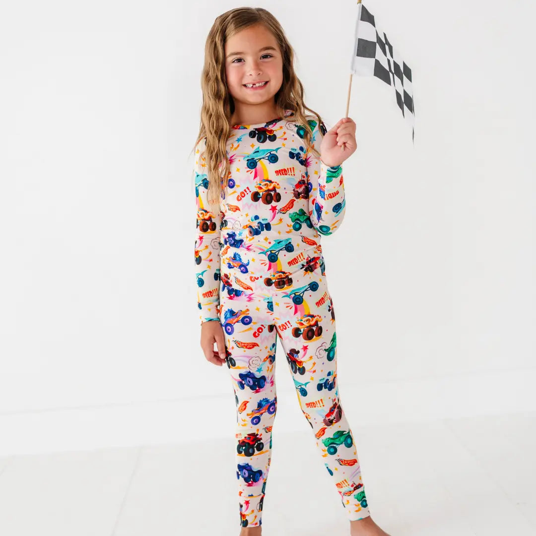 A child holding a checkered racing flag while wearing a cream two-piece pajama set with a colorful monster truck print.