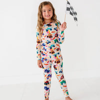 A child holding a checkered racing flag while wearing a cream two-piece pajama set with a colorful monster truck print.
