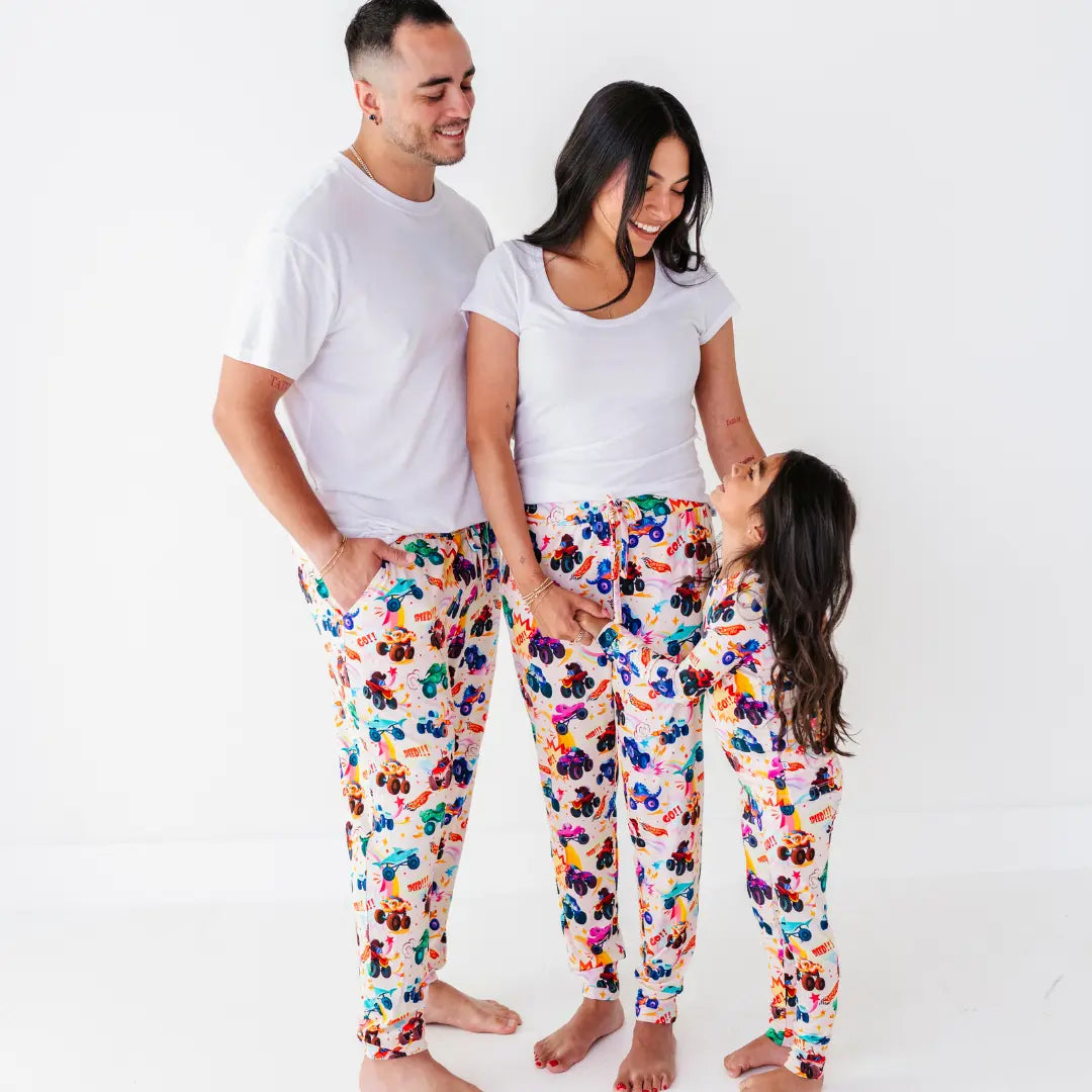A family in white tops and matching monster truck print pajama pants, with a child looking up at the parents.