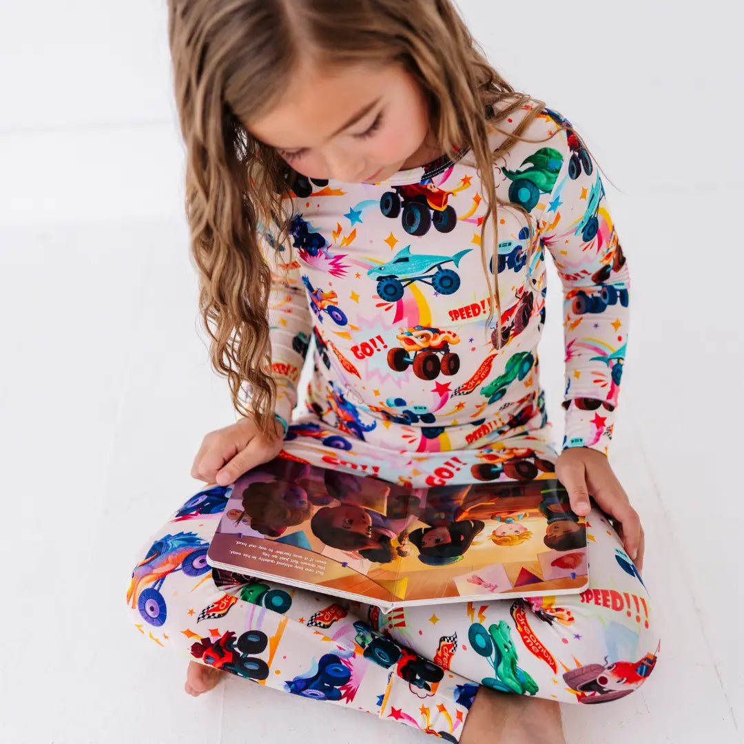 A child sitting cross-legged in cream monster truck print pajamas, reading an open picture book.