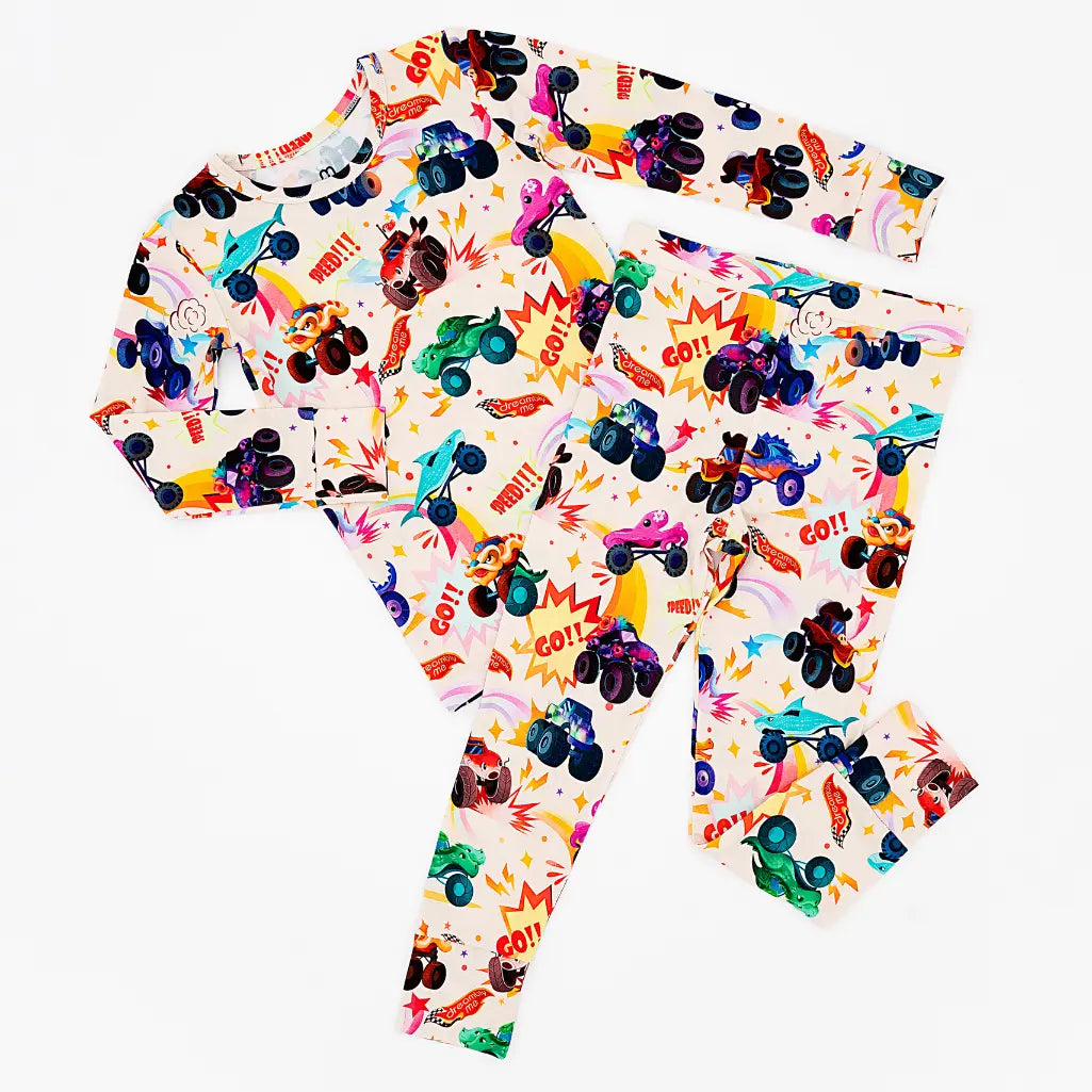 Flat lay of a cream two-piece pajama set with a colorful monster truck print and comic-style bursts on a white background.