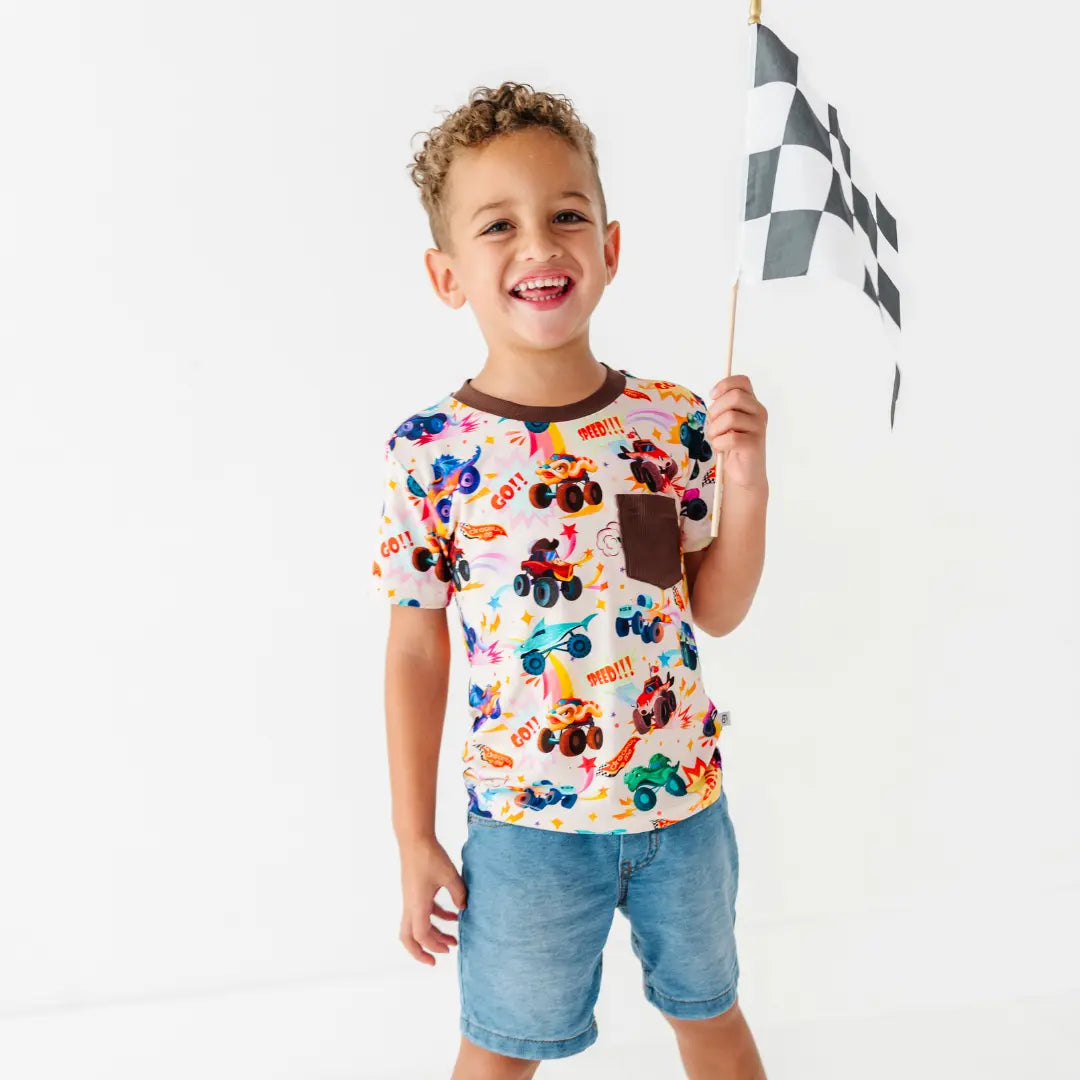 A child holding a checkered racing flag while wearing a monster truck print tee with a brown chest pocket and denim shorts.