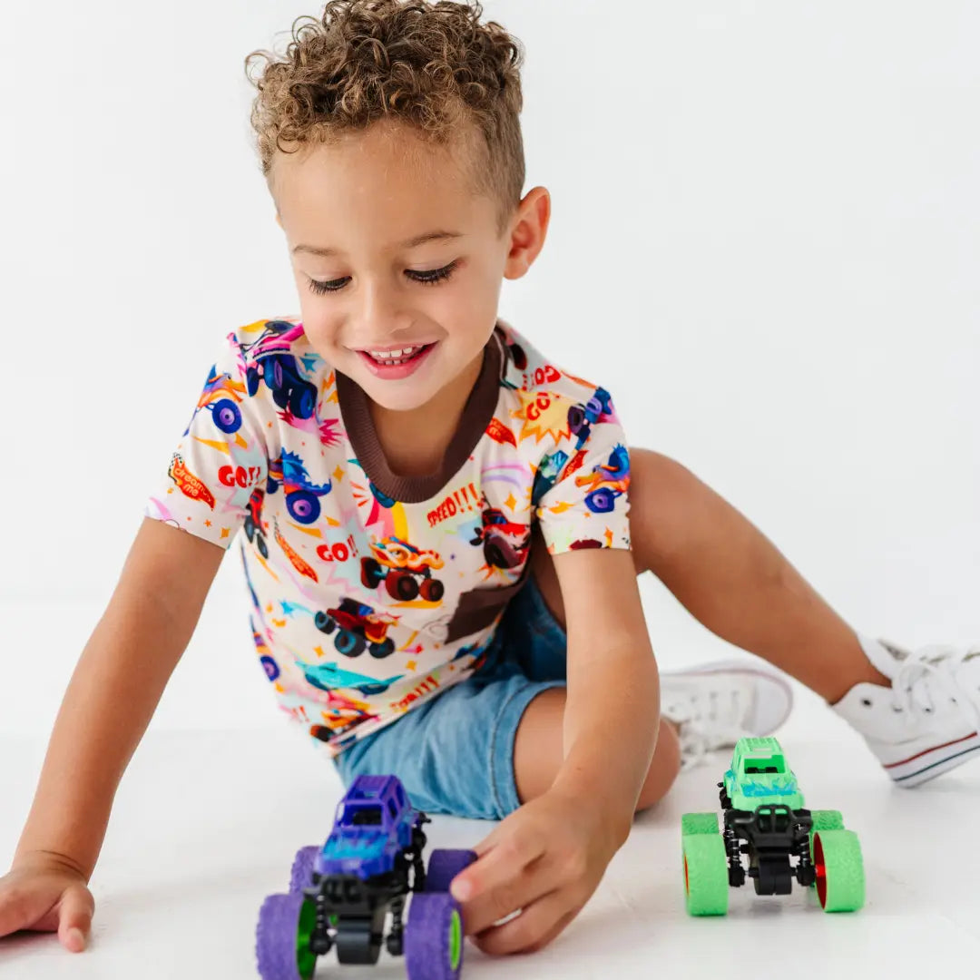 A child in a monster truck print tee and denim shorts playing on the floor with toy monster trucks.