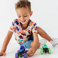A child in a monster truck print tee and denim shorts playing on the floor with toy monster trucks.