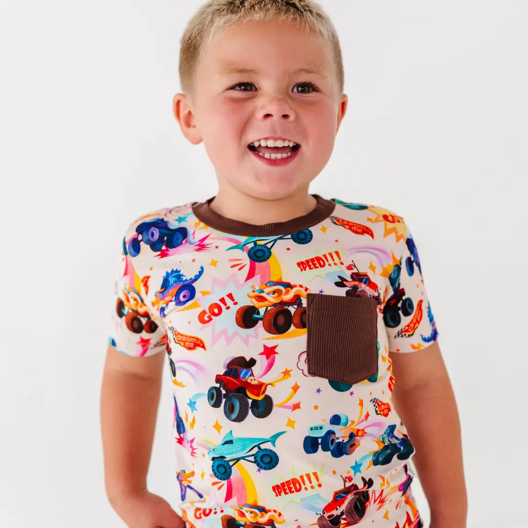 A child laughing in a cream tee with a brown chest pocket and a colorful monster truck print.