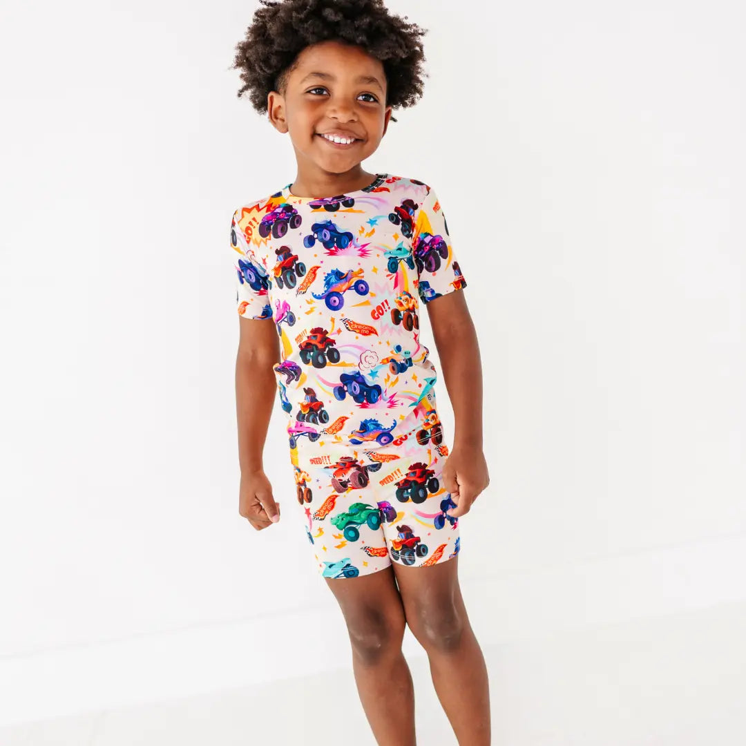 A child smiling in a cream short pajama set with a colorful monster truck print.