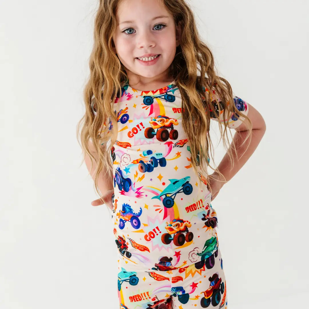 A child posing with hands on hips in a cream short-sleeve pajama set with a colorful monster truck print.