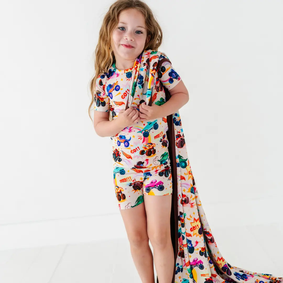 A child in monster truck print shorts pajamas holding a matching print blanket draped over one shoulder.