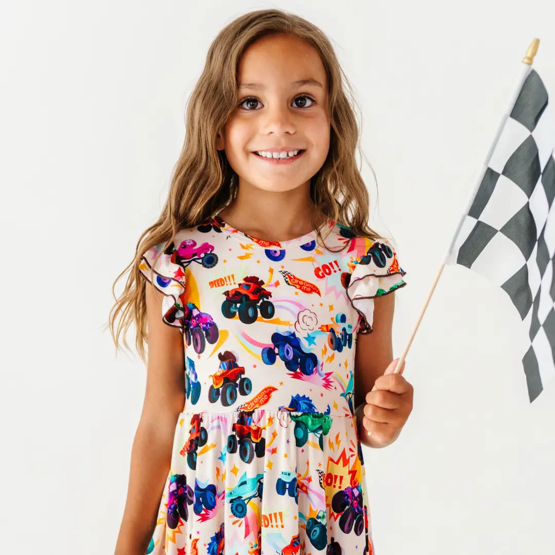 A child smiling and holding a checkered racing flag while wearing a cream flutter-sleeve dress with a monster truck print.