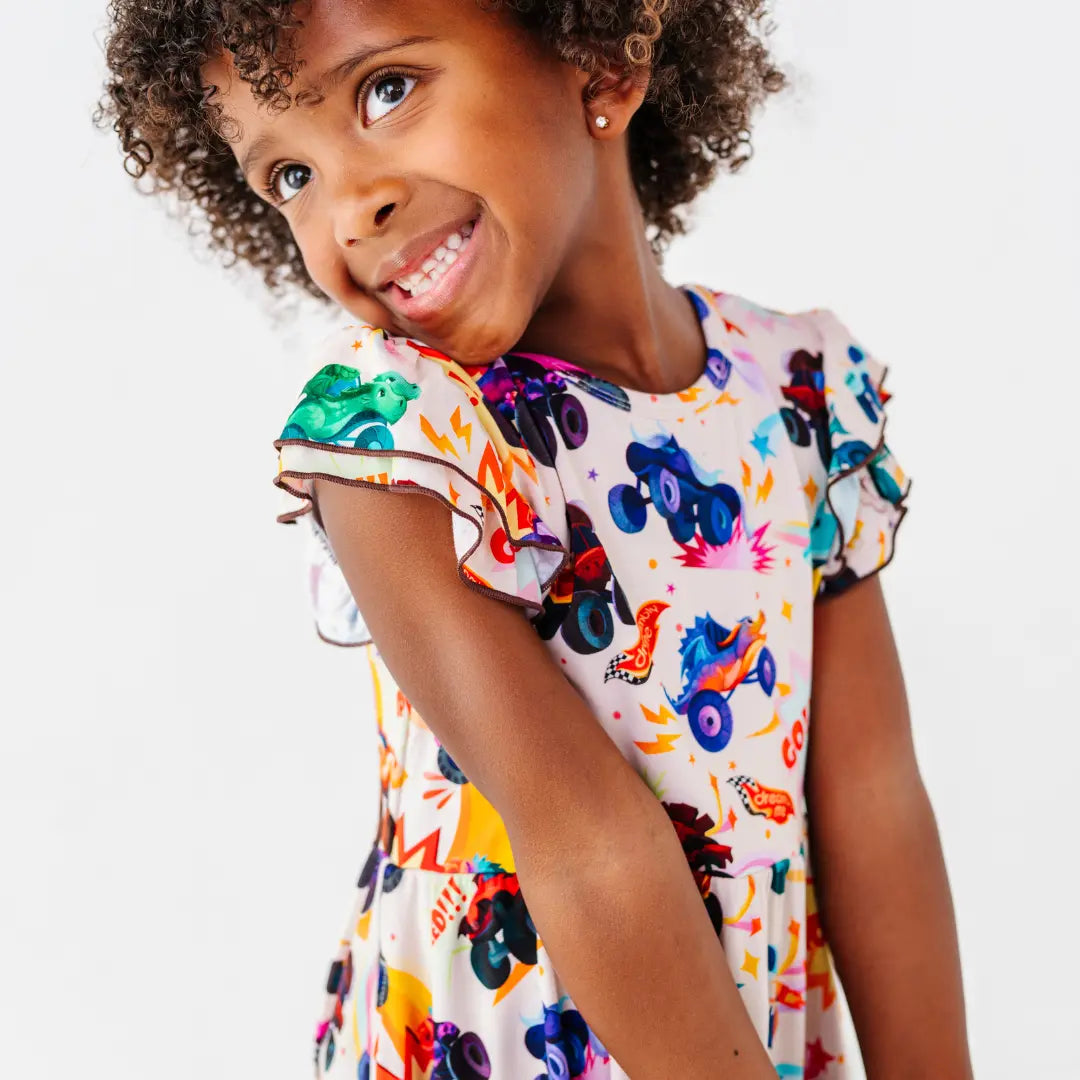 A child smiling over their shoulder in a cream flutter-sleeve dress with a colorful monster truck print.