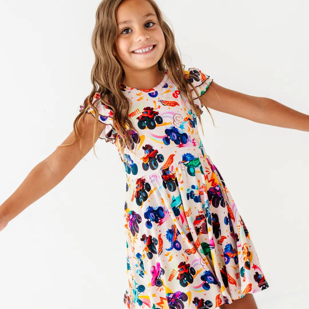 A child with arms outstretched wearing a cream flutter-sleeve twirl dress with a colorful monster truck print.