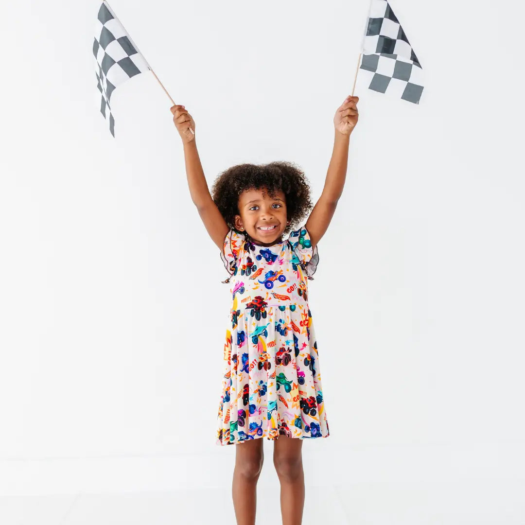 A child raising two checkered racing flags overhead, wearing a cream twirl dress with a colorful monster truck print.