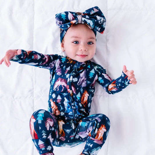 A baby lying on a white quilt and smiling at the camera in a big matching bow and a zip up pajama in a navy print of geometric wolves, triangle pine trees, and snowflakes.
