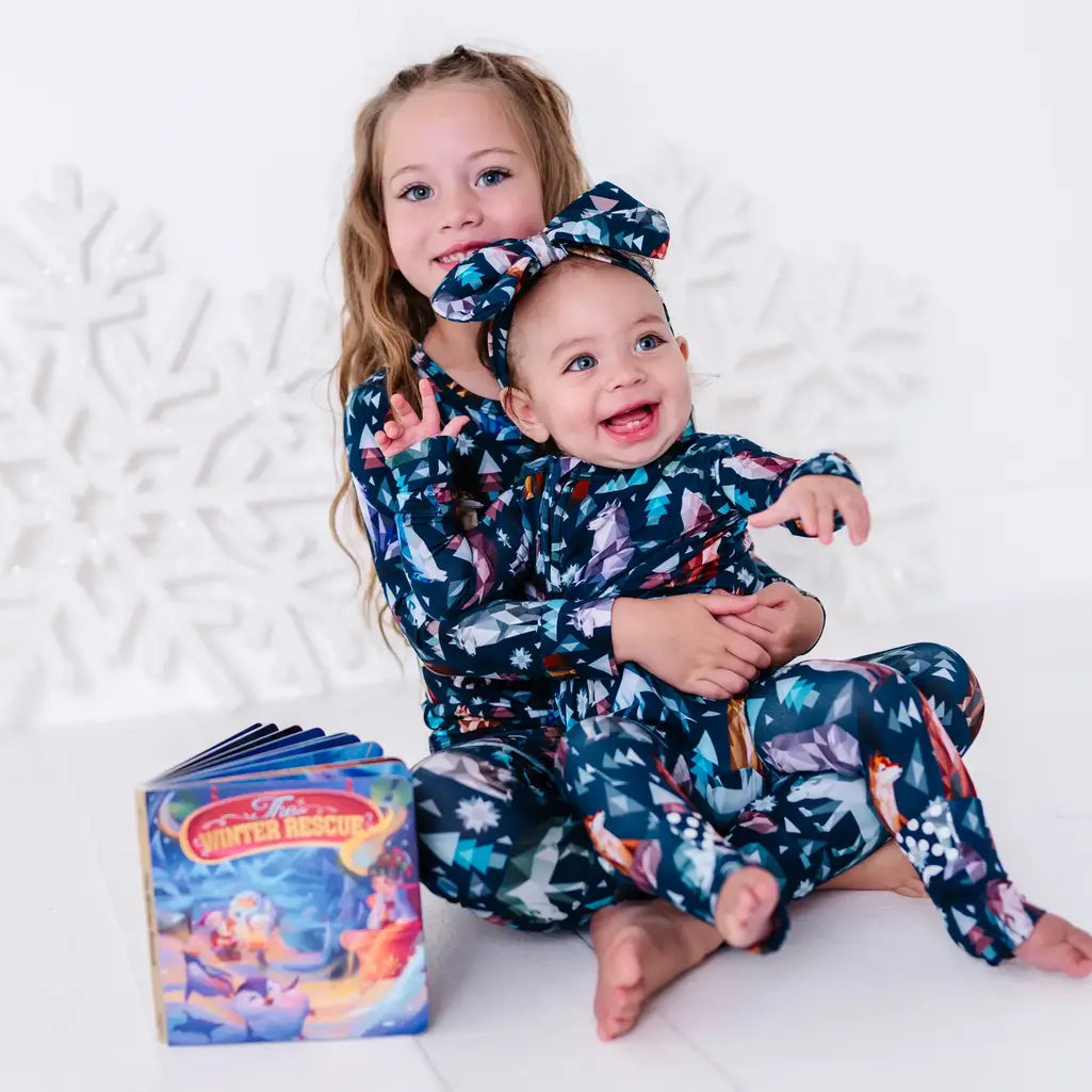 An older sibling holding a laughing baby, in a pajama set and zip up baby pajama with a bow in a navy print of geometric wolves, triangle pine trees, and snowflakes.