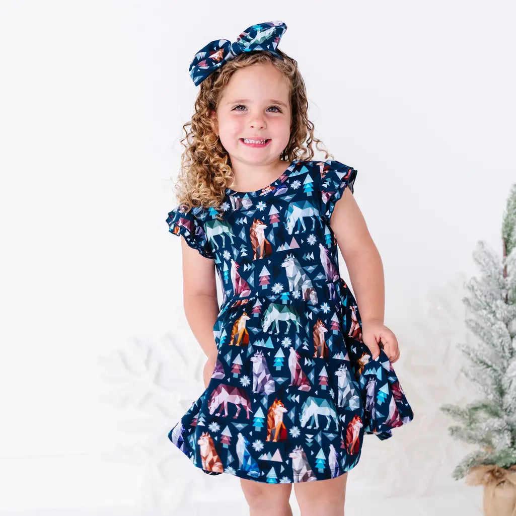 A smiling child holding out the skirt of a flutter sleeve twirl dress with a matching bow headband, in a navy print of geometric wolves, triangle pine trees, and snowflakes.