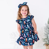 A smiling child holding out the skirt of a flutter sleeve twirl dress with a matching bow headband, in a navy print of geometric wolves, triangle pine trees, and snowflakes.