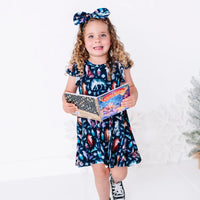 A smiling child holding an open board book at the waist, in a flutter sleeve twirl dress in a navy print of geometric wolves, triangle pine trees, and snowflakes.