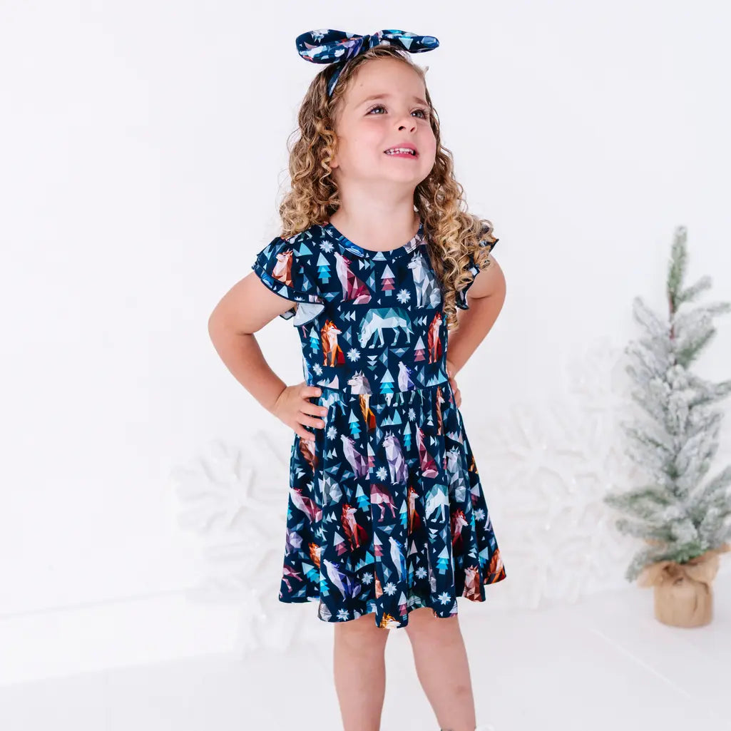 A child standing with hands on the hips and looking up with a smile, in a flutter sleeve twirl dress in a navy print of geometric wolves, triangle pine trees, and snowflakes.
