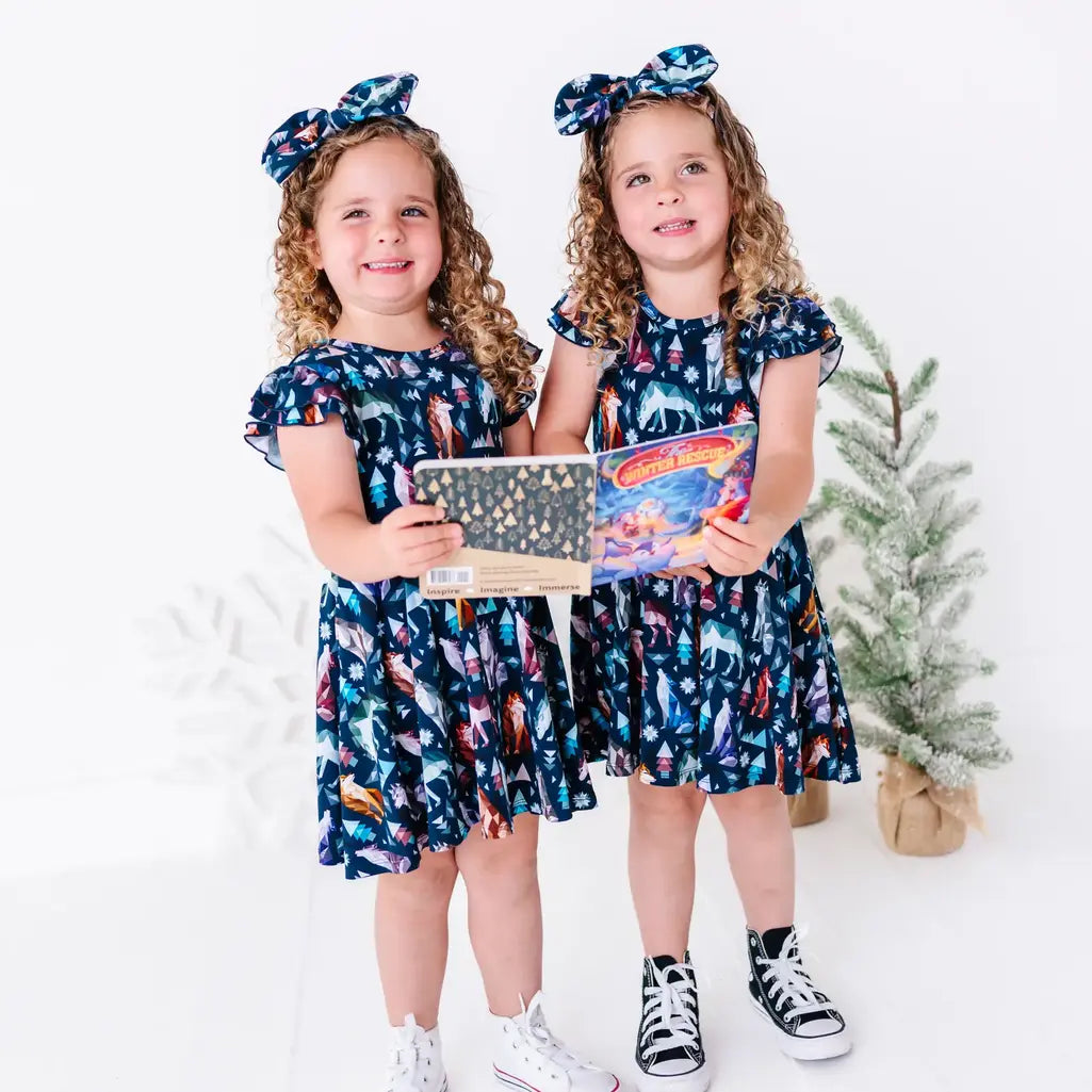 Two kids holding a storybook and smiling in matching twirl dresses in a navy print of geometric wolves, triangle pine trees, and snowflakes.