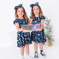 Two kids holding a storybook and smiling in matching twirl dresses in a navy print of geometric wolves, triangle pine trees, and snowflakes.