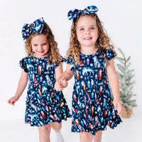 Two kids holding hands and smiling in matching twirl dresses in a navy print of geometric wolves, triangle pine trees, and snowflakes.