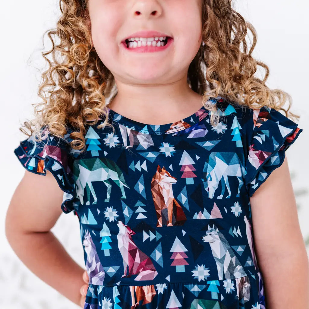 Close up of a child smiling with a hand on the hip, showing a flutter sleeve and bodice of a twirl dress in a navy print of geometric wolves, triangle pine trees, and snowflakes.