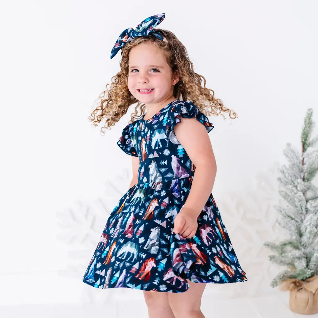 A child twirling with a big smile and the skirt flaring out, in a flutter sleeve dress in a navy print of geometric wolves, triangle pine trees, and snowflakes.