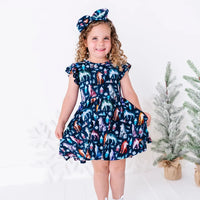 A smiling child holding out one side of the skirt of a flutter sleeve dress with a matching bow headband, in a navy print of geometric wolves, triangle pine trees, and snowflakes.