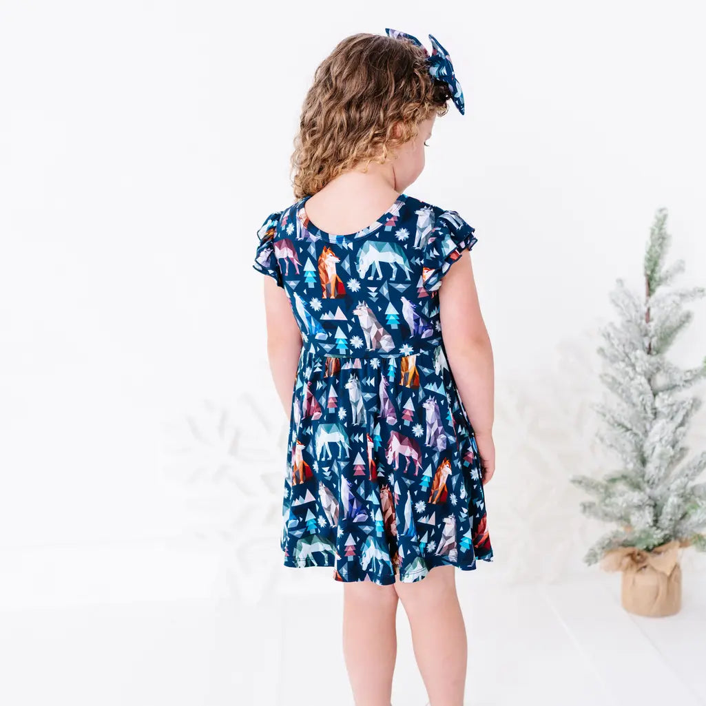 Back view of a child looking down to the side in a flutter sleeve twirl dress with a matching bow headband, in a navy print of geometric wolves, triangle pine trees, and snowflakes.