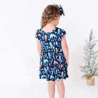 Back view of a child looking down to the side in a flutter sleeve twirl dress with a matching bow headband, in a navy print of geometric wolves, triangle pine trees, and snowflakes.