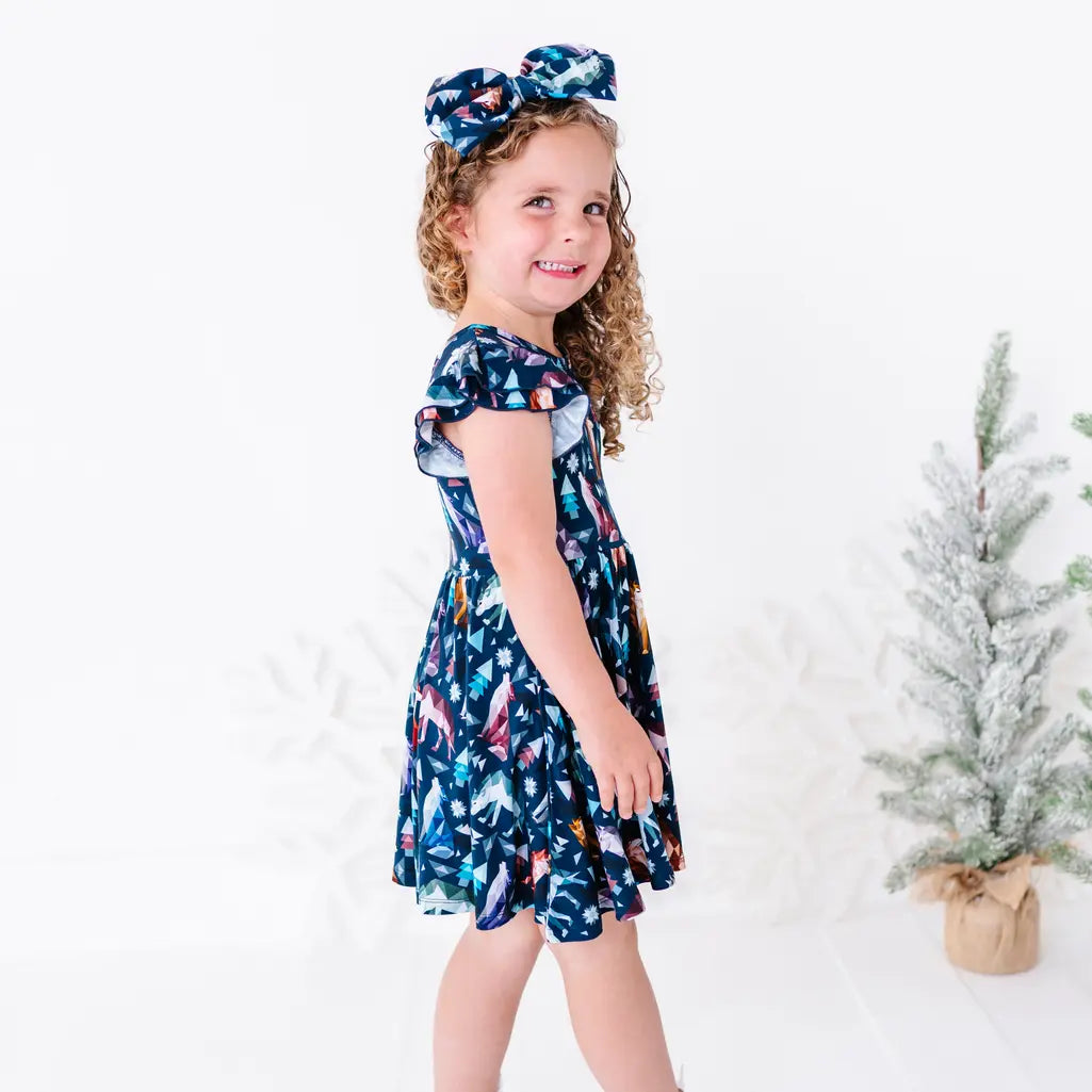 Side view of a child smiling over the shoulder in a flutter sleeve twirl dress with a matching bow headband, in a navy print of geometric wolves, triangle pine trees, and snowflakes.